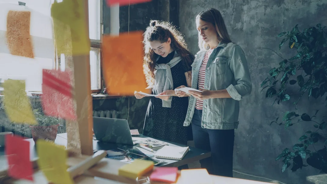 Two women collaborating in a creative workspace with sticky notes and laptop.